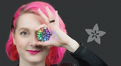 the founder of adafruit holds a circuitboard with many colorful lights up to her eye with the adafruit logo to the side
