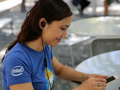 Intel Engineer Indira Negi wearing smart ear buds and looking at her phone while sitting at a table outside